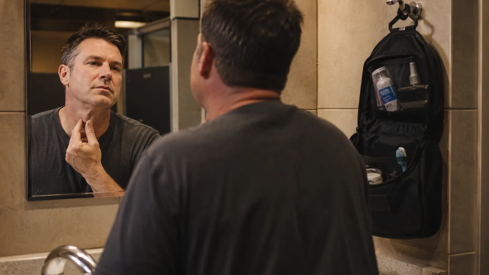 Long-haul trucker standing at a well-lit truck stop bathroom mirror looking sharp in a flannel shirt, hanging toiletry bag on the hook behind him